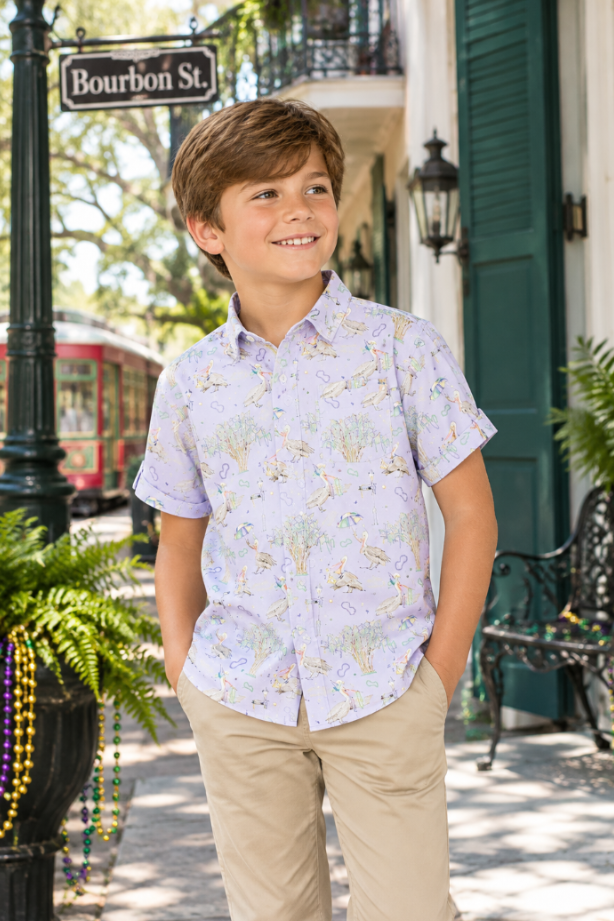 A young boy stands on a street sidewalk wearing a light lavender short-sleeve button-up shirt with a multicolor floral and butterfly print, paired with khaki pants. The background features a green street sign reading 'Bourbon St.